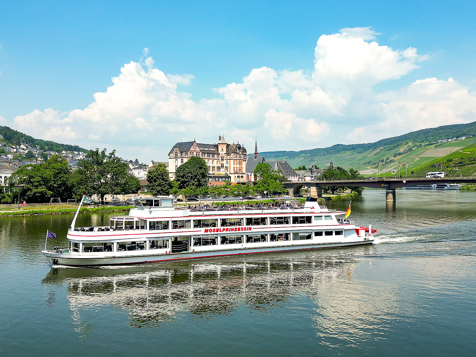 Schifffahrt mit Aussicht auf der Mosel in Bernkastel-Kues Das Schiff ist von vorne seitlich zu sehen. Es fährt moselaufwärts, weg von der Moselbrücke. Im Hintergrund das ehemalige Hotel 3 Könige sowie Weinberge, Häuser und Wälder.