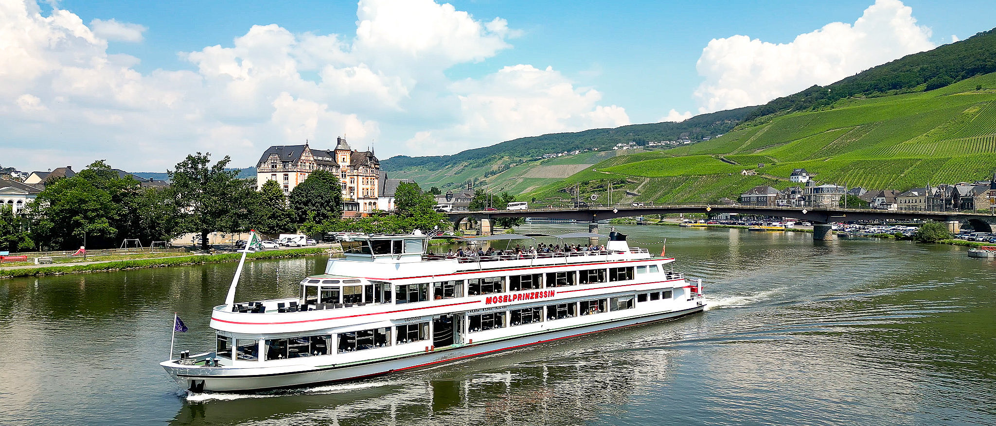 Rundfahrt auf der Mosel in Bernkastel-Kues. Mit Blick auf Weinberge, Moselbrücke und Stadt. Das Schiff fährt moselaufwärts in Richtung der Kamera. Unter blauem Himmel ist die Stadt Bernkastel-Kues Kulisse, außerdem noch Weinberge und Wälder.